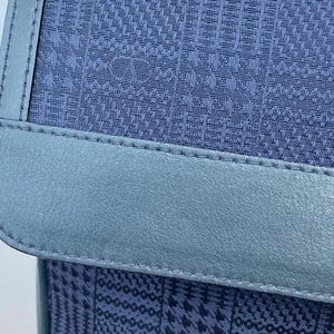 May include: Close-up of a navy blue handbag with a textured pattern. The bag features a light blue leather trim and stitching. The bag's design includes a repeating pattern of the letter "V".