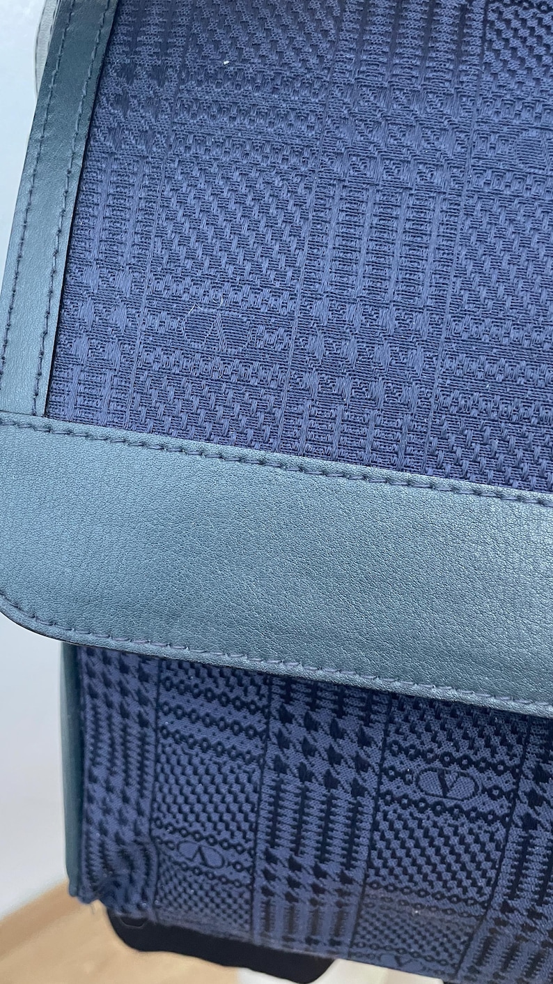 May include: Close-up of a navy blue handbag with a textured pattern. The bag features a light blue leather trim and stitching. The bag's design includes a repeating pattern of the letter "V".