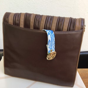 May include: A brown leather clutch with a pleated top section in alternating shades of brown and tan. The clutch features a blue and white patterned strap and a gold-toned clasp. The bag is sitting on a white box.