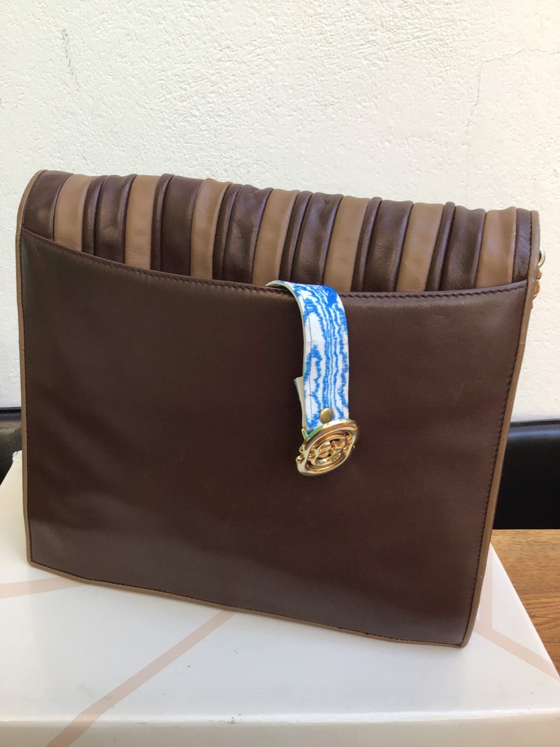May include: A brown leather clutch with a pleated top section in alternating shades of brown and tan. The clutch features a blue and white patterned strap and a gold-toned clasp. The bag is sitting on a white box.