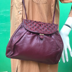 Könnte beinhalten: Burgunderfarbene Leder-Umhängetasche mit geflochtenem Überschlag und goldfarbenem Verschluss. Die Tasche hat eine runde Form und einen langen, dünnen Riemen. Die Tasche wird auf einer Schaufensterpuppe mit beigem Hemd präsentiert.