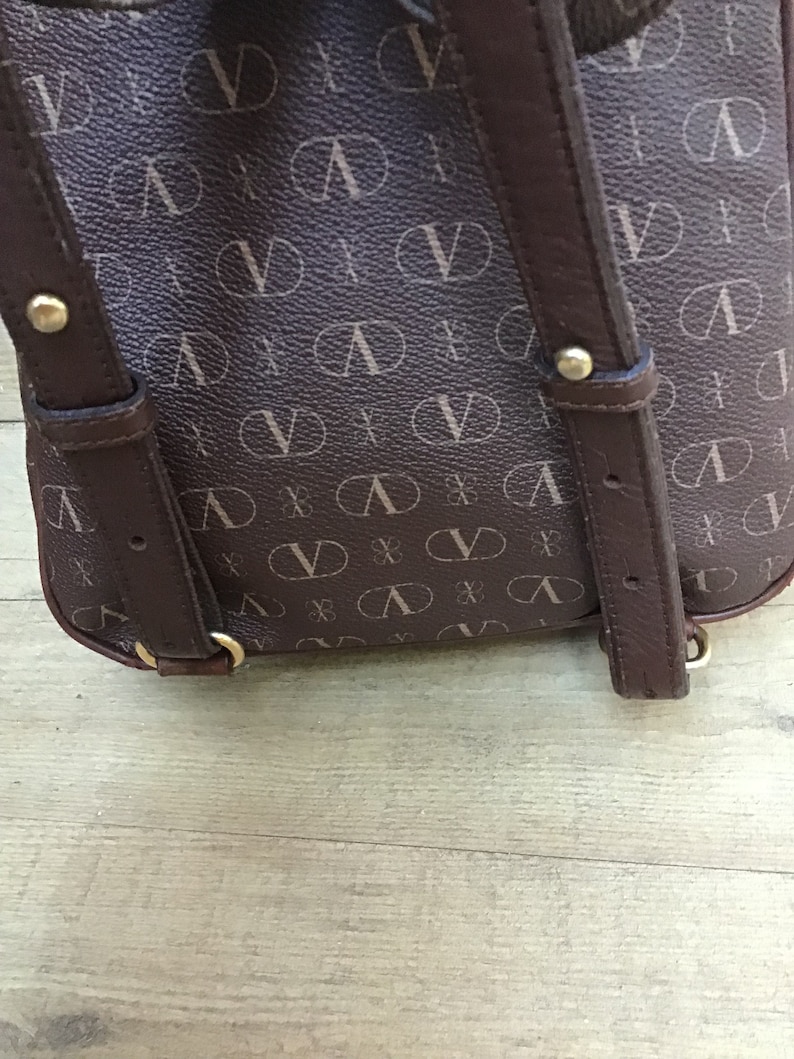 K&ouml;nnte beinhalten: Ein brauner Lederrucksack mit verstellbaren Tr&auml;gern und einem sich wiederholenden Muster des Buchstabens "V" auf der Vorderseite.