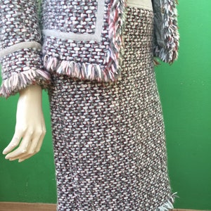 Peut inclure: Un tailleur en tweed à deux pièces avec un motif tissé gris, blanc et bordeaux. La veste a une bordure à franges et des empiècements en daim. La jupe est mi-longue avec une frange assortie.