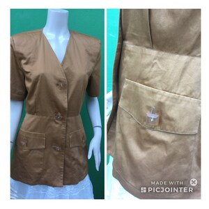May include: A tan-coloured jacket with short sleeves, a V-neck, and three decorative buttons. It has two front pockets and a fitted waist. The right side shows a close-up of a pocket with a decorative button.