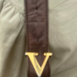 May include: Close-up of a brown leather strap with a gold-colored metal V-shaped buckle. The strap appears to be part of a bag or accessory, set against a light beige background.