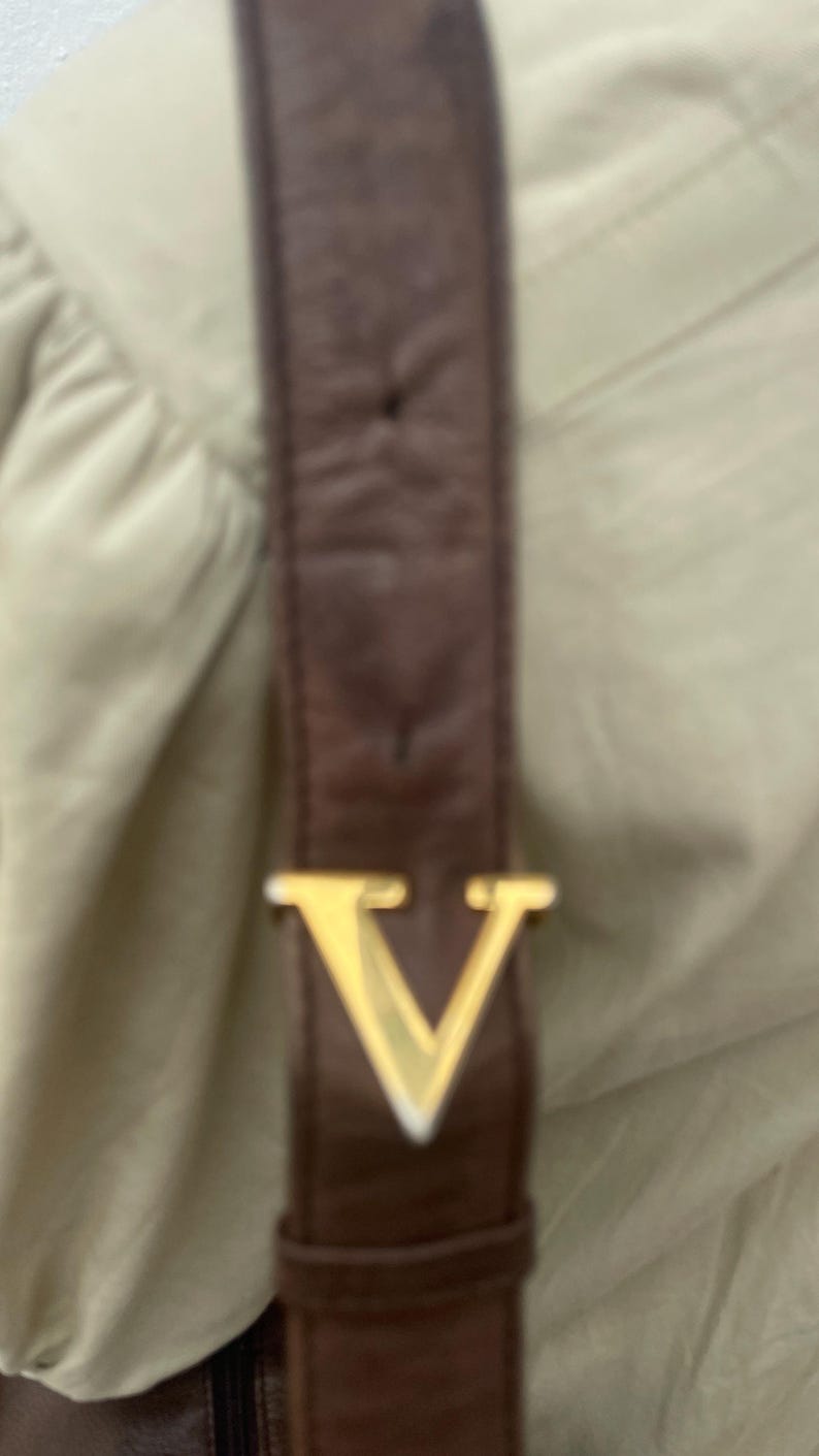 May include: Close-up of a brown leather strap with a gold-colored metal V-shaped buckle. The strap appears to be part of a bag or accessory, set against a light beige background.