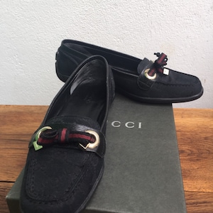Peut inclure: Mocassins Gucci noirs avec un détail de nœud et des accessoires dorés. Les chaussures sont présentées sur une boîte Gucci. Les mocassins ont un design classique, adapté à diverses occasions.