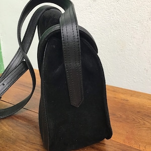 May include: A black suede handbag with a leather handle and shoulder strap. The bag has a rectangular shape with a curved top flap. The leather handle is stitched and attached to the bag. The bag is sitting on a wooden surface.