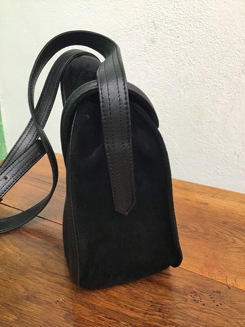 May include: A black suede handbag with a leather handle and shoulder strap. The bag has a rectangular shape with a curved top flap. The leather handle is stitched and attached to the bag. The bag is sitting on a wooden surface.
