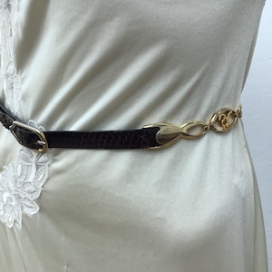 Peut inclure: Une ceinture marron texturée imitation peau de serpent avec une boucle et un fermoir dorés. La ceinture est portée sur un vêtement crème avec des détails de dentelle blanche. La ceinture a une chaîne dorée décorative.