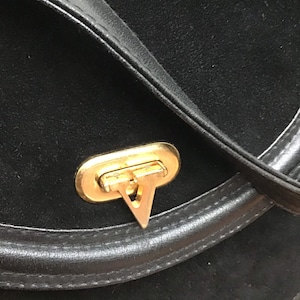 May include: Black suede shoulder bag with a gold-tone clasp. The bag features a rounded shape with a leather trim and a flap closure. The clasp has a distinctive V-shaped design.