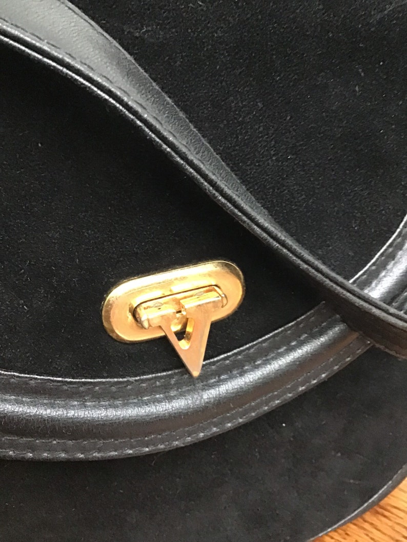 May include: Black suede shoulder bag with a gold-tone clasp. The bag features a rounded shape with a leather trim and a flap closure. The clasp has a distinctive V-shaped design.