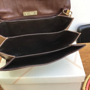 May include: Brown leather Valentino Garavani handbag with a gold-tone clasp and logo. The bag has multiple interior compartments and is open, revealing the dark brown interior. The bag is sitting on a white box.