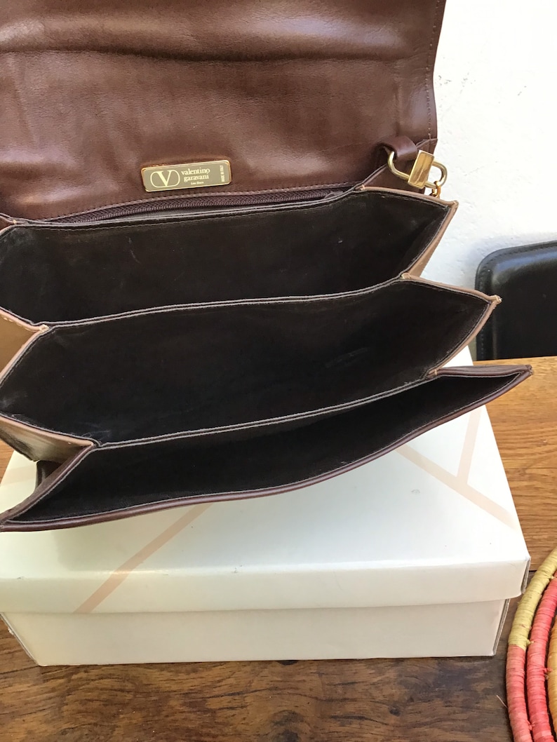 May include: Brown leather Valentino Garavani handbag with a gold-tone clasp and logo. The bag has multiple interior compartments and is open, revealing the dark brown interior. The bag is sitting on a white box.