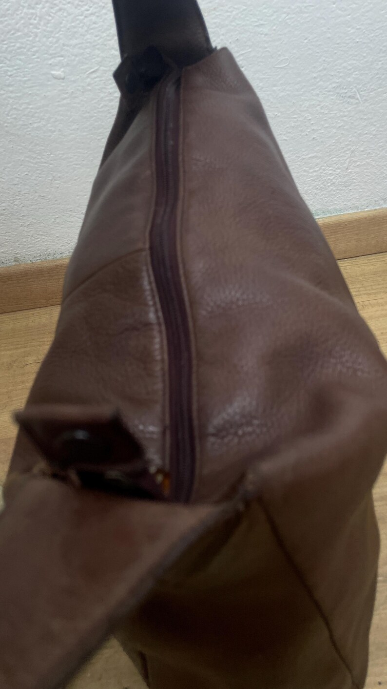 May include: A brown leather shoulder bag with a zipper closure. The bag has a flap with a buckle. The leather has a textured appearance. The bag is designed to be worn over the shoulder.