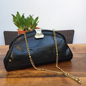 Puede incluir: Bolso de mano negro con una correa de cadena dorada. El bolso tiene un patrón texturizado, similar a la piel de serpiente, en la parte delantera y un cierre plateado. La cadena es de color dorado delicado. El bolso está sobre una superficie de madera.