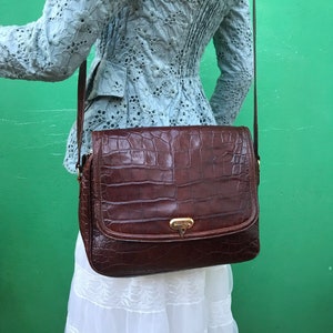 Op de afbeelding: Een bruine leren crossbodytas met een krokodillenstructuur. De tas heeft een rechthoekige vorm met een klepsluiting en een goudkleurige sluiting. De tas wordt gedragen over een lichtblauwe jas en witte rok.
