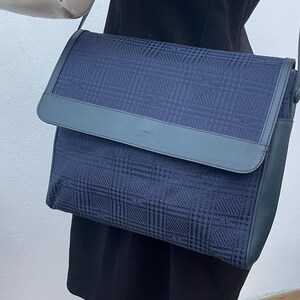 May include: A navy blue crossbody bag with a plaid pattern. The bag features a flap closure and a matching shoulder strap. The bag is made of a textured fabric and has a leather trim.