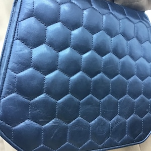 May include: A navy blue shoulder bag with a quilted, honeycomb pattern. The bag has a flap closure and a strap. The stitching is visible, and the bag appears to be made of leather or a similar material. The bag is a dark blue colour.
