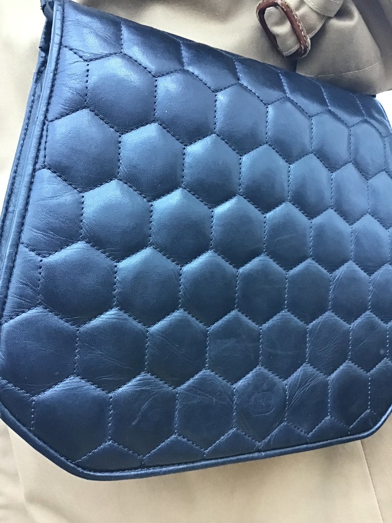 May include: A navy blue shoulder bag with a quilted, honeycomb pattern. The bag has a flap closure and a strap. The stitching is visible, and the bag appears to be made of leather or a similar material. The bag is a dark blue colour.