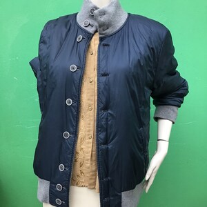 Può includere: Giacca trapuntata blu navy con colletto e polsini grigi, abbottonata. La giacca è sovrapposta a una camicia beige abbottonata. Il manichino indossa una gonna bianca. Lo sfondo è verde.
