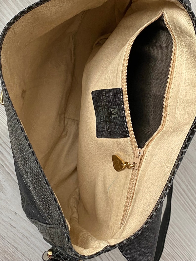 May include: The inside of a black leather handbag with a zippered pocket and a gold-colored metal tag that reads "Made in Italy" and "Murano Venice".