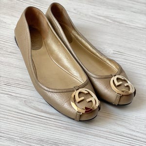 K&ouml;nnte beinhalten: Ein Paar goldene Ballerinas aus Leder mit einem goldenen, ineinander verschlungenen G-Logo an der Spitze.
