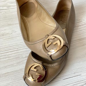 K&ouml;nnte beinhalten: Ein Paar goldene Gucci Ballerinas aus Leder mit einem goldenen, ineinandergreifenden G-Logo an der Spitze. Die Schuhe befinden sich auf einer hellbraunen Holzoberfl&auml;che.