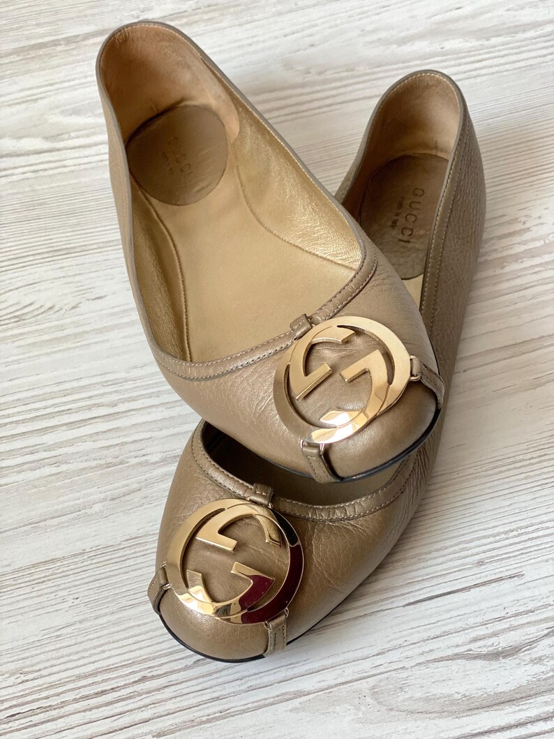 K&ouml;nnte beinhalten: Ein Paar goldene Gucci Ballerinas aus Leder mit einem goldenen, ineinandergreifenden G-Logo an der Spitze. Die Schuhe befinden sich auf einer hellbraunen Holzoberfl&auml;che.