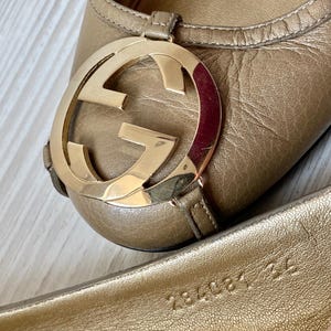 K&ouml;nnte beinhalten: Ein Paar goldene Ballerinas aus Leder mit einem gro&szlig;en, goldenen, ineinandergreifenden GG-Logo an der Spitze. Die Ballerinas haben ein Gr&ouml;&szlig;enetikett auf der Innensohle mit der Aufschrift "234081 422".