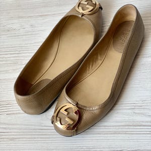 K&ouml;nnte beinhalten: Ein Paar braune Gucci Ballerinas aus Leder mit goldenen, ineinandergreifenden G-Logo-Akzenten.