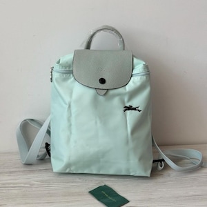 Puede incluir: Una mochila azul claro con cierre de solapa y asa superior. La solapa es de un tono azul más claro y presenta un botón negro. Un logotipo negro de un caballo corriendo está en la parte delantera. Incluye correas para los hombros y una pequeña etiqueta verde.