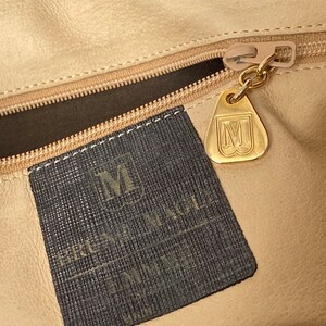 May include: A black leather handbag with a gold zipper and a black fabric label with the text "BRUNO MAGLI" and a gold metal tag with the letter "M".