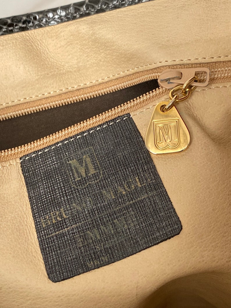 May include: A black leather handbag with a gold zipper and a black fabric label with the text "BRUNO MAGLI" and a gold metal tag with the letter "M".