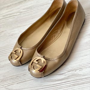 K&ouml;nnte beinhalten: Ein Paar goldene Ballerinas aus Leder mit einem goldenen, ineinandergreifenden G-Logo an der Spitze.
