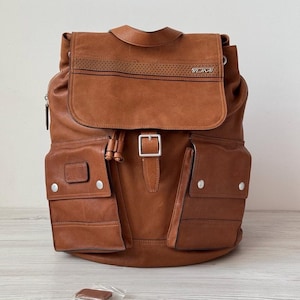 May include: A brown leather backpack with multiple pockets and a flap closure. The backpack features a silver buckle and the brand name "TUMI" on the flap. The bag has two side pockets with silver button closures and a drawstring closure. A small, brown leather tag is in front of the bag.