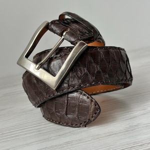 Peut inclure: Une ceinture en cuir marron foncé avec une boucle argentée. La ceinture a une surface texturée et à motifs, enroulée sur elle-même. La boucle est rectangulaire et brillante. La ceinture est sur une surface en bois clair.