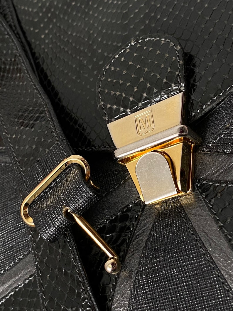 May include: A black leather handbag with a gold-toned clasp and a snakeskin pattern. The clasp has a metal plate with the letter "M" on it.