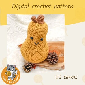 May include: A crocheted amigurumi gourd with a smiling face, brown stem, and a yellow-brown color. The gourd is sitting on a wooden surface with pine cones.
