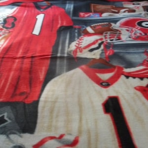 Puede incluir: Una manta de forro polar roja y blanca con temática de fútbol americano. La manta presenta una camiseta de fútbol con el número "1" en ella, un casco de fútbol y un par de tacos de fútbol. La manta también tiene las palabras "Bulldogs" y "Georgia" en ella.
