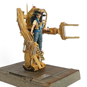 May include: A detailed figurine of a character operating a yellow and black exoskeleton. The figure is standing on a gray platform. The exoskeleton has a claw-like arm and a red light on top. The character is wearing blue pants and a tan shirt.