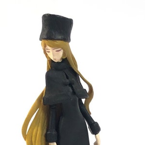May include: A black and gold figurine of a woman wearing a black coat and a black hat. She is holding a black briefcase.