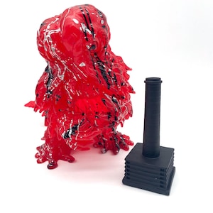 May include: A red, abstract sculpture with black and silver paint splatters. Next to it is a black, cylindrical object on a tiered, square base. The sculpture has a textured, organic form.