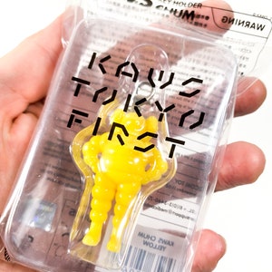 Peut inclure: Porte-clés en plastique jaune avec une figurine stylisée et le texte "KAWS TOKYO FIRST" sur l'emballage.