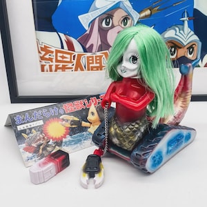 Op de afbeelding: Een vintage speelgoed met een zeemeermin met groen haar en een rode top, gemonteerd op een blauw-zwarte tankrupsbasis. Een kleine kaart met Japanse tekst en twee extra speelgoedcomponenten zijn ook aanwezig. Een ingelijste poster staat op de achtergrond.