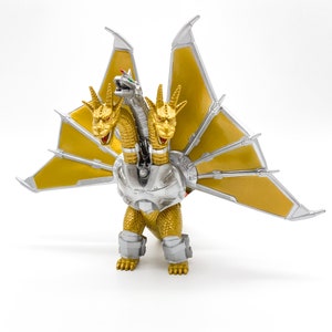 Peut inclure: Une figurine d'action dorée et argentée d'un monstre à trois têtes avec des ailes. Le monstre a une poitrine en argent métallique et des jambes et des ailes dorées.