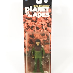 Puede incluir: Una figura de acción de plástico de Lucius, un personaje de la franquicia El Planeta de los Simios. La figura es verde y marrón y está empaquetada en una blíster de plástico transparente. El empaquetado presenta las palabras "Ultra Detail Figure" y "El Planeta de los Simios".