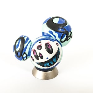 Può includere: Una scultura stravagante con un volto da cartone animato, più occhi e un ampio sorriso. Le orecchie sono blu e azzurre con dettagli neri. La scultura è montata su una base color argento. Il design è giocoso e colorato.