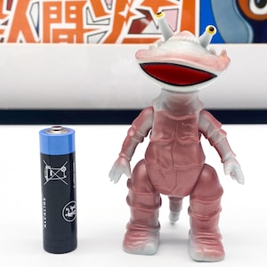 Peut inclure: Une figurine de jouet rose et blanche avec une grande bouche et deux antennes. La figurine a une surface texturée et se tient sur un fond blanc.