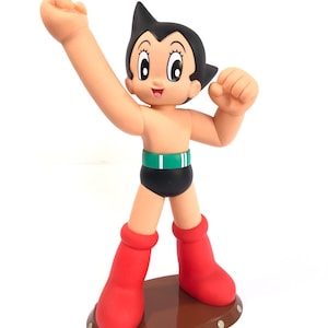 May include: A red and black Astro Boy figurine with a green belt and a brown base. The figurine is standing with its right arm raised and its left fist clenched.